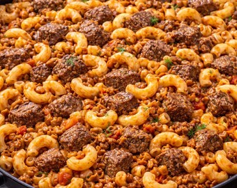 May include: A close-up shot of a large pan filled with a hearty meal. The dish features elbow macaroni, ground meat, and chunks of meat, all mixed in a tomato-based sauce. Sprigs of green herbs are scattered throughout.