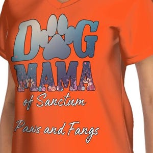 May include: Orange V-neck t-shirt with the text "Dog Mama of Sanctum Paws and Fangs" in a blue and floral design. The shirt has short sleeves and is made of a soft material.