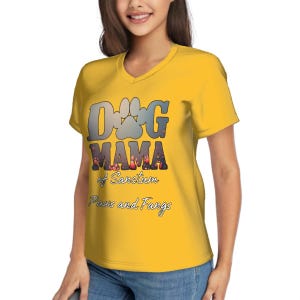 May include: A yellow V-neck t-shirt with the text "Dog Mama" in a stylized font, featuring paw prints and the words "of Sanctum, Paws and Fangs". The shirt is made of a soft material.