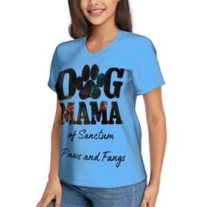 May include: A light blue V-neck t-shirt with the words "DOG MAMA" in black, with a paw print design. Below the words are the phrases "of Sanctum" and "Paws and Fangs" in black script.
