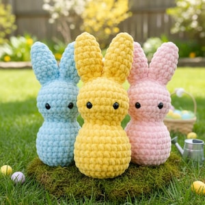 May include: Three crochet bunny figures in pastel colors: light blue, yellow, and pink. Each bunny has black button eyes and upright ears. They are displayed on a bed of green moss, suggesting an Easter theme.