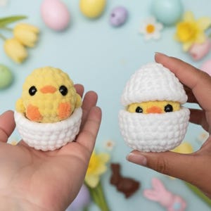 May include: Two handmade crochet Easter chicks, one fully hatched and the other partially hatched from white crochet eggshells. The chicks are yellow with orange beaks and feet, and black eyes. The background includes Easter eggs and flowers.