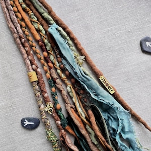 May include: A collection of handmade dreadlocks in various shades of brown, orange, and green, adorned with fabric, beads, and metal accents. Some dreads feature a light blue fabric. The dreadlocks are arranged on a gray linen surface.