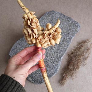 Puede incluir: Una varita de madera hecha a mano con un acabado natural, adornada con detalles tallados y un grupo de elementos beige. El mango está envuelto con hilo de colores. La varita se sostiene en una mano.
