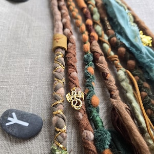 Peut inclure: Gros plan sur des dreadlocks faits à la main dans diverses teintes terreuses, notamment marron, vert et orange. Les dreads sont ornées d'accents et de breloques dorés, et sont disposées sur une surface en lin neutre. Deux pierres grises avec des marques blanches sont en arrière-plan.