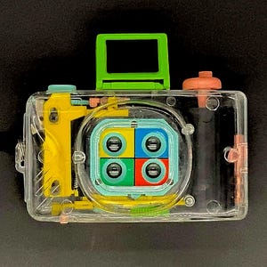 May include: A clear plastic toy camera with a green viewfinder, yellow internal components, and a colorful lens array. The camera has a black strap and a pink button. The design is a playful take on a classic camera.