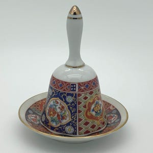 Peut inclure: Une cloche décorative en céramique avec une poignée blanche et des accents dorés, posée sur une soucoupe assortie. La cloche présente un motif floral et géométrique coloré en rouge, bleu et or. La soucoupe a un bord doré.
