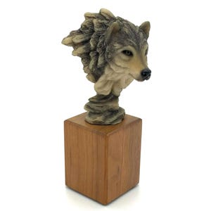May include: A detailed sculpture of a wolf's head, featuring realistic fur textures and facial details. The wolf is mounted on a square, wooden base. The sculpture's colors include shades of gray, brown, and beige.