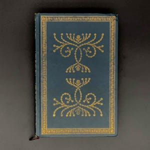 May include: A hardcover book with a dark teal cover and gold decorative filigree. The cover features a symmetrical design of floral and starburst patterns. The edges of the cover are also trimmed in gold. A red ribbon bookmark is visible.