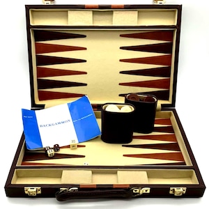 Può includere: Un set da backgammon aperto con custodia marrone e superficie di gioco color crema con punte triangolari marroni e beige. Il set include due bicchieri per dadi marrone scuro, pedine bianche e marroni, due dadi, un cubo del raddoppio e un libretto blu.