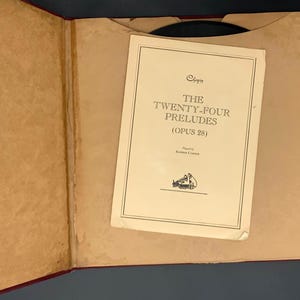 Op de afbeelding: Open boek met een crèmekleurige pagina getiteld "The Twenty-Four Preludes (Opus 28)". Het boek is open op een vakje met de pagina. De kaft van het boek is donkerrood en de pagina's zijn lichtbruin.