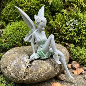 Puede incluir: Estatua de hada gris sentada sobre una piedra. El hada lleva una falda verde claro y un top a juego. Las alas son grandes y detalladas. La estatua tiene un rostro y cabello detallados.
