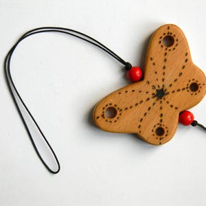 Può includere: Un ciondolo in legno a forma di farfalla con un cordino nero. Il ciondolo è marrone chiaro con linee tratteggiate e un disegno a stella. Due perline rosse sono attaccate al ciondolo. Il cordino è nero.