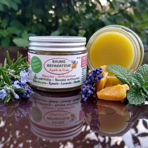 Puede incluir: Un frasco de vidrio de 50 ml con bálsamo amarillo y tapa plateada, etiquetado como Baume Reparateur. La etiqueta incluye texto como Muscles - Articulations, Loatifolia e ingredientes como romero, lavanda y menta. Hierbas frescas y cera al lado.