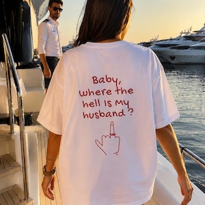 Puede incluir: Una camiseta blanca de manga corta con texto en rojo en la espalda que dice: "Baby, where the hell is my husband?", acompañada de un gráfico de líneas rojas de una mano con un anillo en el dedo medio, en un corte casual y relajado.