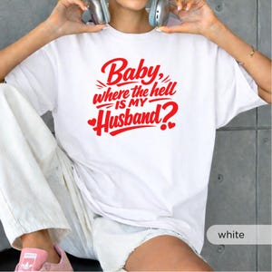 以下が含まれることがあります： 赤い文字で「Baby, where the hell is my Husband?」とハートのアクセントが入った白いTシャツ。カジュアルなスタイルのシャツです。