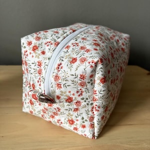 May include: A rectangular floral print cosmetic bag with a white zipper. The bag features a pattern of red flowers and green leaves on a white background. The bag is sitting on a wooden surface.