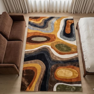 May include: A rectangular area rug with an abstract design in shades of brown, gold, black, and gray. The rug features a layered, organic pattern reminiscent of a topographic map. The rug is placed on a light-colored wooden floor.