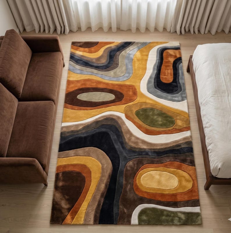 May include: A rectangular area rug with an abstract design in shades of brown, gold, black, and gray. The rug features a layered, organic pattern reminiscent of a topographic map. The rug is placed on a light-colored wooden floor.