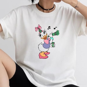 May include: A white short-sleeved t-shirt featuring a graphic print of a cartoon duck wearing a pink polka-dot party hat, a purple shirt, and pink shoes. The duck is holding a green party horn and a small green cup, with black musical notes.