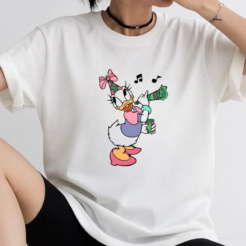 May include: A white short-sleeved t-shirt featuring a graphic print of a cartoon duck wearing a pink polka-dot party hat, a purple shirt, and pink shoes. The duck is holding a green party horn and a small green cup, with black musical notes.