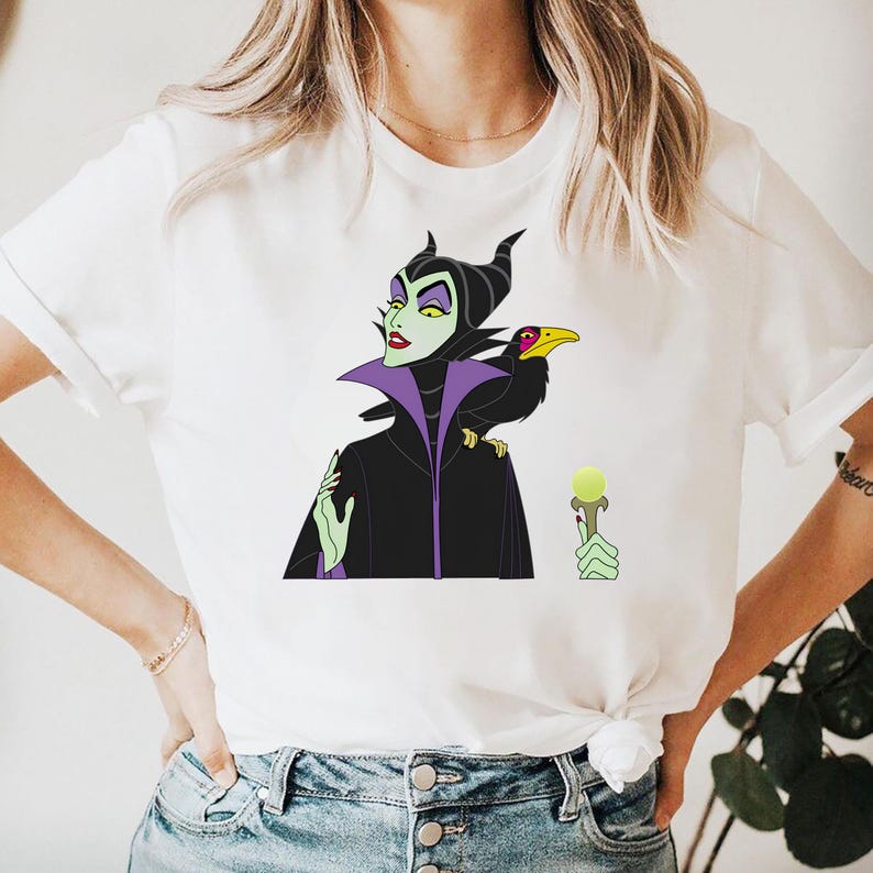 May include: White t-shirt featuring a cartoon illustration of a character with green skin, black horns, and a black cloak. A black raven sits on the character's shoulder. The character is holding a staff with a green orb.