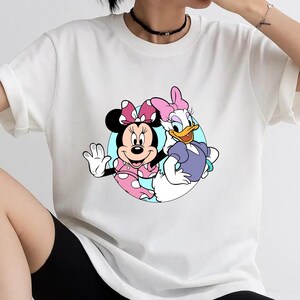 May include: A white short-sleeved t-shirt featuring a circular graphic print of Minnie Mouse and Daisy Duck. Minnie Mouse wears a pink polka-dot dress and bow, while Daisy Duck wears a purple top and a pink bow, set against a light blue background.