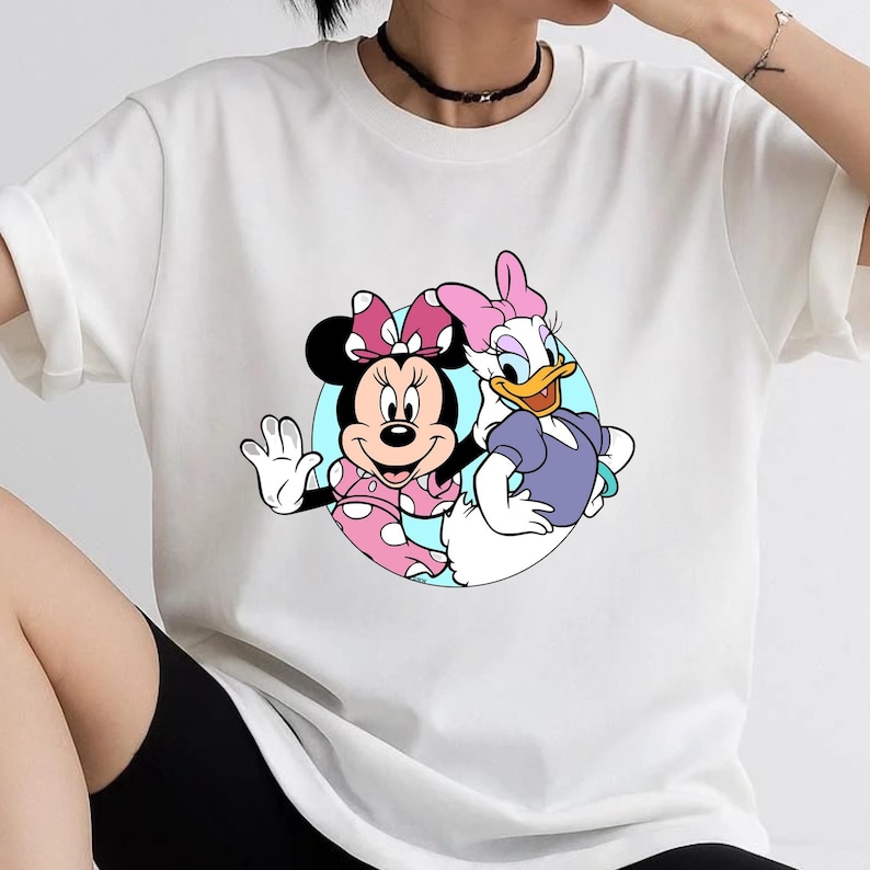 May include: A white short-sleeved t-shirt featuring a circular graphic print of Minnie Mouse and Daisy Duck. Minnie Mouse wears a pink polka-dot dress and bow, while Daisy Duck wears a purple top and a pink bow, set against a light blue background.