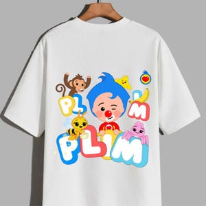 May include: A white short-sleeved t-shirt featuring a colourful graphic print of the cartoon character Plim Plim with a blue hairstyle, a red nose, and a red shirt, surrounded by a monkey, a bee, a yellow bird, a banana, and the text PLIM PLIM.