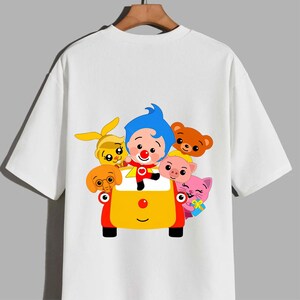 May include: A white short-sleeved t-shirt featuring a colourful graphic print of a cartoon character with blue hair and a red suit standing in a yellow and red car, surrounded by a yellow rabbit, brown bear, pink pig, pink cat, and orange elephant.