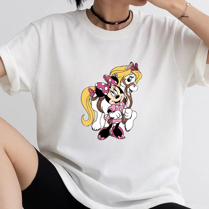 May include: A white short-sleeved t-shirt featuring a graphic print of Minnie Mouse with a pink polka-dot bow and dress, standing next to a white pony with a long blonde mane and a pink bow. The design is centered on the front of the shirt.