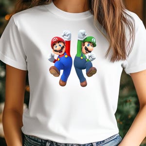 Op de afbeelding: Wit T-shirt met een afbeelding van Mario en Luigi, personages uit de Super Mario-videogameserie. Mario is in rood en blauw, en Luigi is in groen en blauw. Beiden springen met hun vuisten in de lucht.