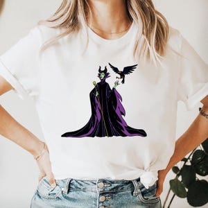 May include: White t-shirt featuring a cartoon illustration of a character in a black and purple gown, holding a staff, with a black bird perched on their hand. The character has green skin and black horns.