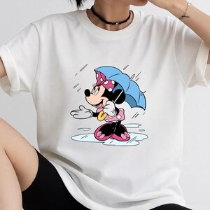 May include: A white short-sleeved t-shirt featuring a graphic print of a cartoon mouse character wearing a pink polka-dot dress and bow, holding a light blue umbrella while standing in a puddle during a rain shower with falling raindrops.
