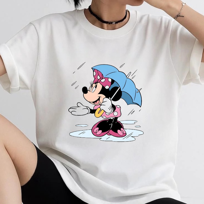 May include: A white short-sleeved t-shirt featuring a graphic print of a cartoon mouse character wearing a pink polka-dot dress and bow, holding a light blue umbrella while standing in a puddle during a rain shower with falling raindrops.