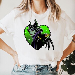 May include: White t-shirt featuring a graphic of a green-skinned character with black horns and a black bird. The character has purple accents and is set against a green and white background. The bird has a yellow eye and beak.