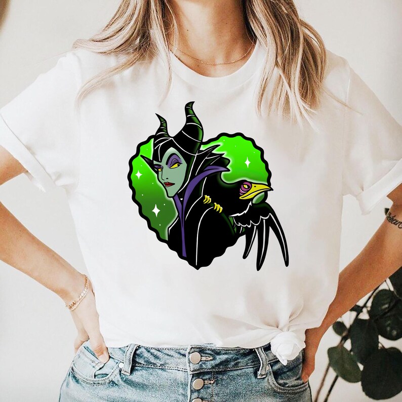 May include: White t-shirt featuring a graphic of a green-skinned character with black horns and a black bird. The character has purple accents and is set against a green and white background. The bird has a yellow eye and beak.