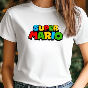 Op de afbeelding: Wit T-shirt met de tekst "SUPER MARIO" in een kleurrijk, blokkerig lettertype. De tekst is zwart omlijnd, met de letters in blauw, geel, rood en groen. Het shirt heeft een klassieke ronde hals.