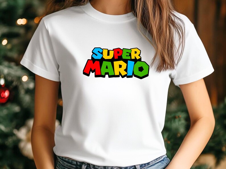 Op de afbeelding: Wit T-shirt met de tekst "SUPER MARIO" in een kleurrijk, blokkerig lettertype. De tekst is zwart omlijnd, met de letters in blauw, geel, rood en groen. Het shirt heeft een klassieke ronde hals.