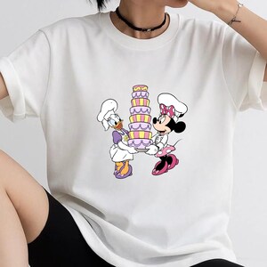 May include: A white short-sleeved t-shirt featuring a graphic print of Daisy Duck and Minnie Mouse wearing chef hats and aprons. They are holding a tall, multi-layered cake with pink, yellow, and purple stripes between them on the front of the shirt.