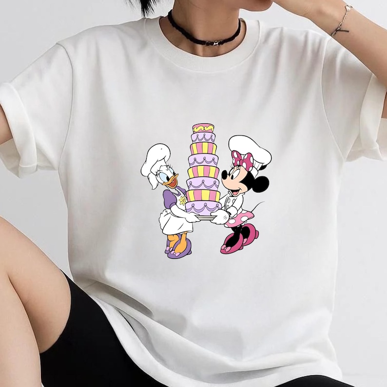 May include: A white short-sleeved t-shirt featuring a graphic print of Daisy Duck and Minnie Mouse wearing chef hats and aprons. They are holding a tall, multi-layered cake with pink, yellow, and purple stripes between them on the front of the shirt.