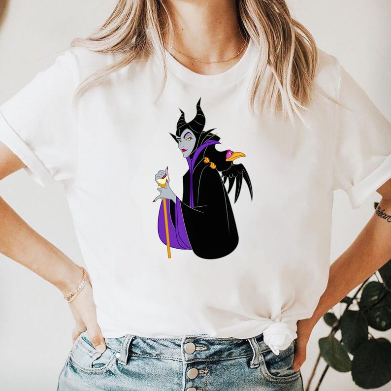 May include: White t-shirt featuring a cartoon illustration of a character with black horns, a black cloak with purple accents, and a raven on the shoulder. The character is holding a staff. The design is centered on the shirt.