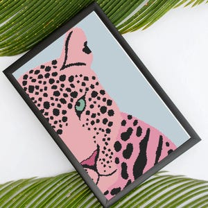May include: A framed art print featuring a pink leopard with black spots and stripes, set against a light blue background. The leopard's eye is a striking teal color. The artwork is in a black frame, creating a modern aesthetic.