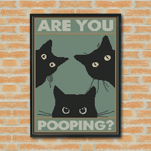 May include: A framed cross-stitch wall art piece featuring three black cat silhouettes on a sage green background. The text ARE YOU POOPING? is printed in a block font at the top and bottom. The cats have visible eyes and whiskers.