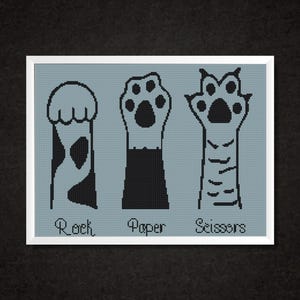 May include: A framed cross-stitch design featuring three stylized cat paws representing "Rock, Paper, Scissors." The paws are black on a light blue background, with the words below each paw. The frame is white.