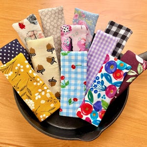 May include: A collection of rectangular fabric pan handle covers in various patterns including yellow floral, acorn print, blue gingham with cherries, purple plaid, black and white check, and colorful floral designs, all resting inside a black cast iron skillet.