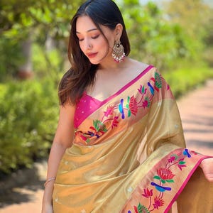 May include: A gold-colored sari with a pink border and floral embroidery. The sari features a pink blouse and is draped over the shoulders. The embroidery includes colorful birds and flowers.