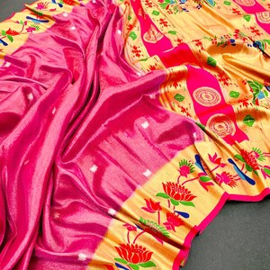 May include: A vibrant pink saree with a shimmering, silky texture. The border features a gold design with floral and bird motifs in red, green, and blue. The saree is draped, showcasing its flowing fabric.