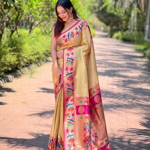 May include: A beige saree with intricate floral and bird embroidery in pink, blue, and green. The saree features a vibrant pink border and a decorative fringe. The woman is wearing the saree outdoors.