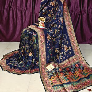 May include: A navy blue sari with a detailed floral and animal print design. The sari features a decorative border with pink, orange, and green accents. A small figurine sits on the draped fabric.